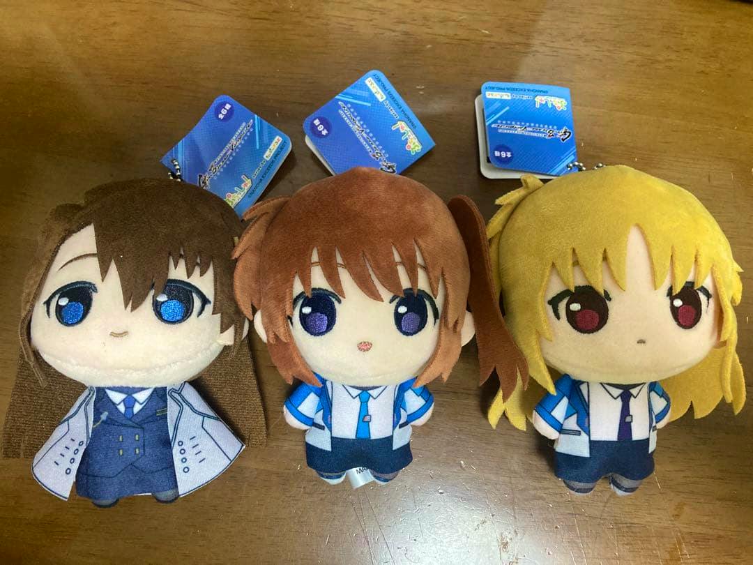 Three plush toys of Nanoha, Fate, and Hayate arranged side by side on a wooden surface.