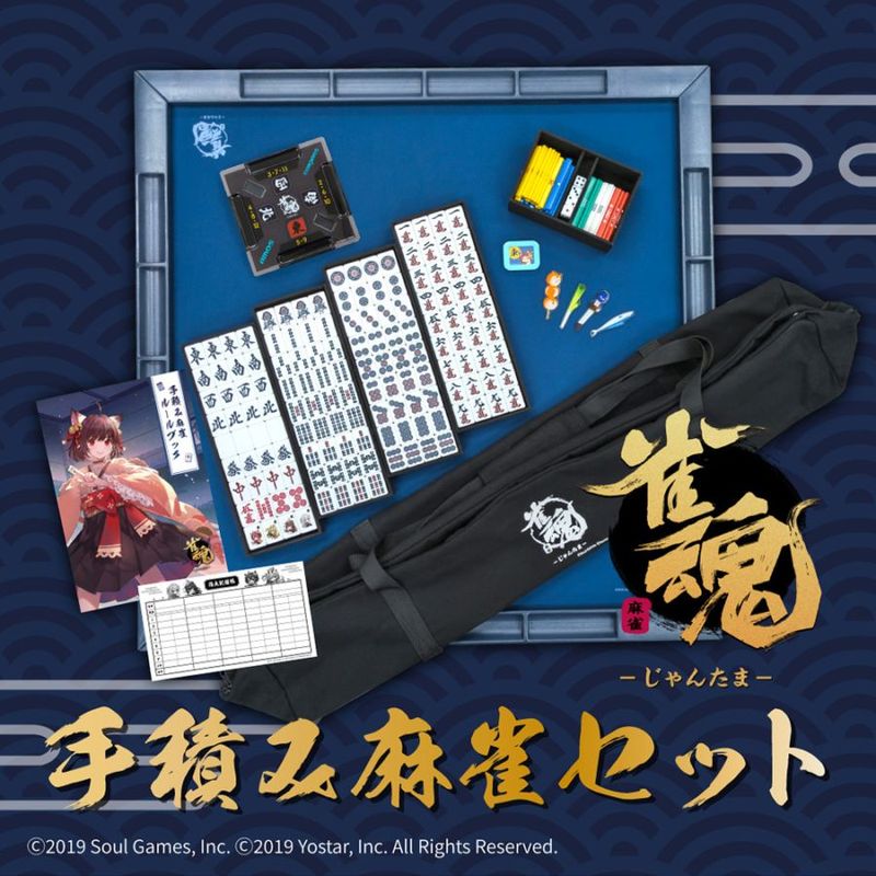 Mahjong set components arranged on a blue tray: tiles, racks, scoring sticks, markers, score sheet, character card, and a black carrying bag with a gold logo.