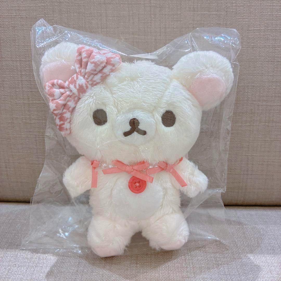Front view of the Maison de FLEUR Korilakkuma plush bag charm with a pink patterned bow and pink button, shown inside clear plastic packaging.