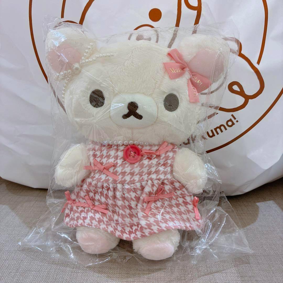 White Korilakkuma plush wearing a pink houndstooth dress with ribbon bows and a pearl necklace, shown inside clear plastic packaging.