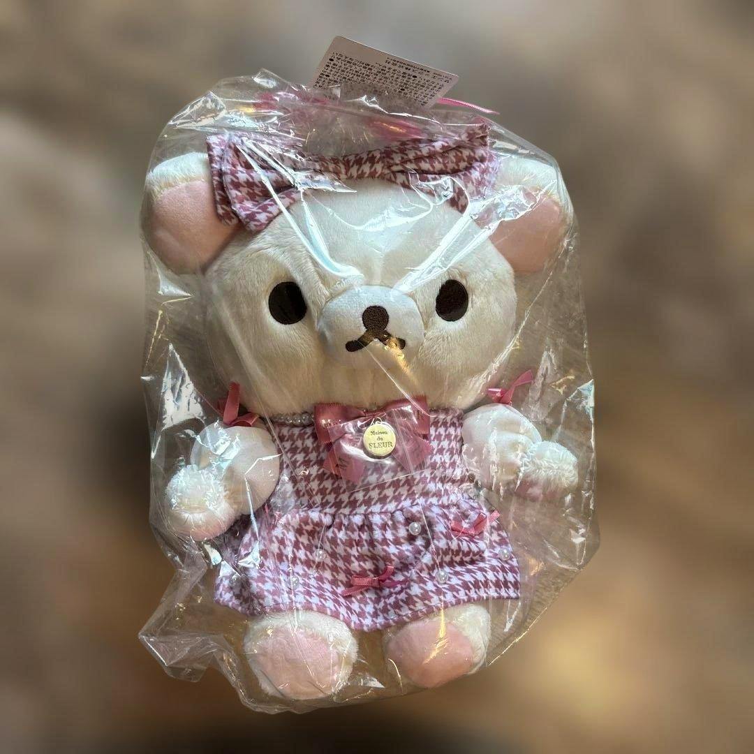 Rilakkuma plush wearing a pink houndstooth dress and bow, shown inside clear plastic packaging.