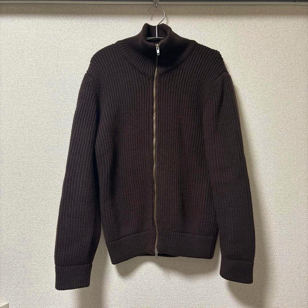 Front view of a ribbed zip-up driver's knit sweater hanging on a hanger against a wall.