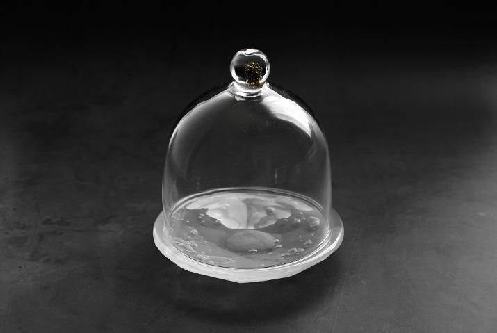Clear glass bell-shaped dome with a rounded knob on top sitting on a shallow glass base against a dark background.