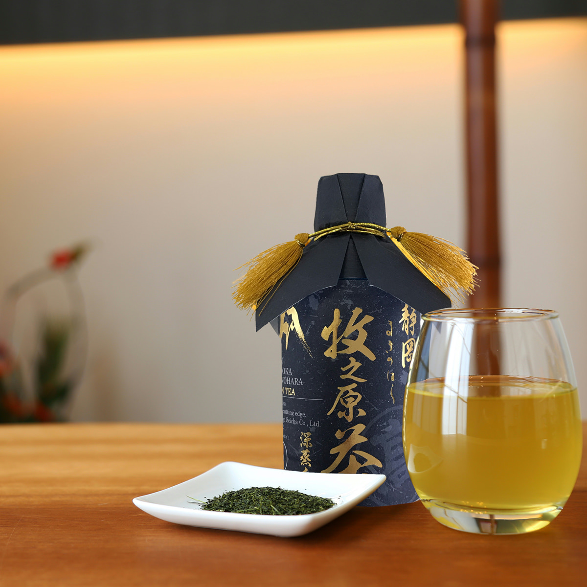 A wrapped bottled green tea next to a glass of brewed tea and a small dish of loose tea leaves on a wooden surface.
