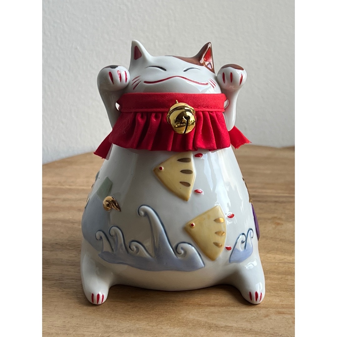 Front view of a ceramic maneki-neko piggy bank figure wearing a red ruffled collar with a gold bell, photographed on a wooden surface.