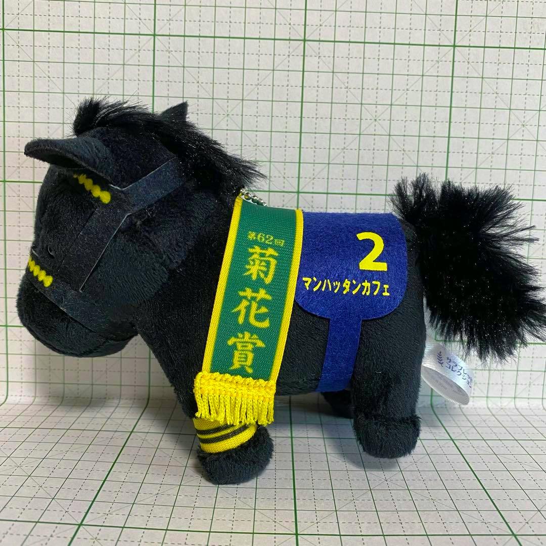 Black plush horse mascot keychain (マンハッタンカフェ) with a green-and-yellow sash and a blue saddle marked "2" shown on a grid background.