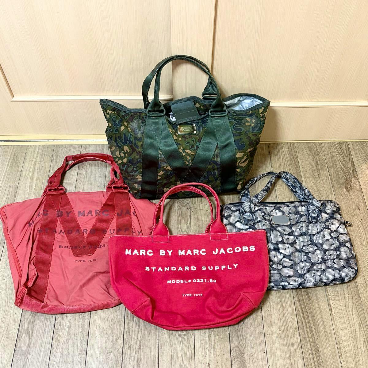 Four Marc by Marc Jacobs bags arranged on a wooden floor: a green camouflage tote, a bright red tote with white "MARC BY MARC JACOBS" text, a faded red tote, and a gray patterned bag.