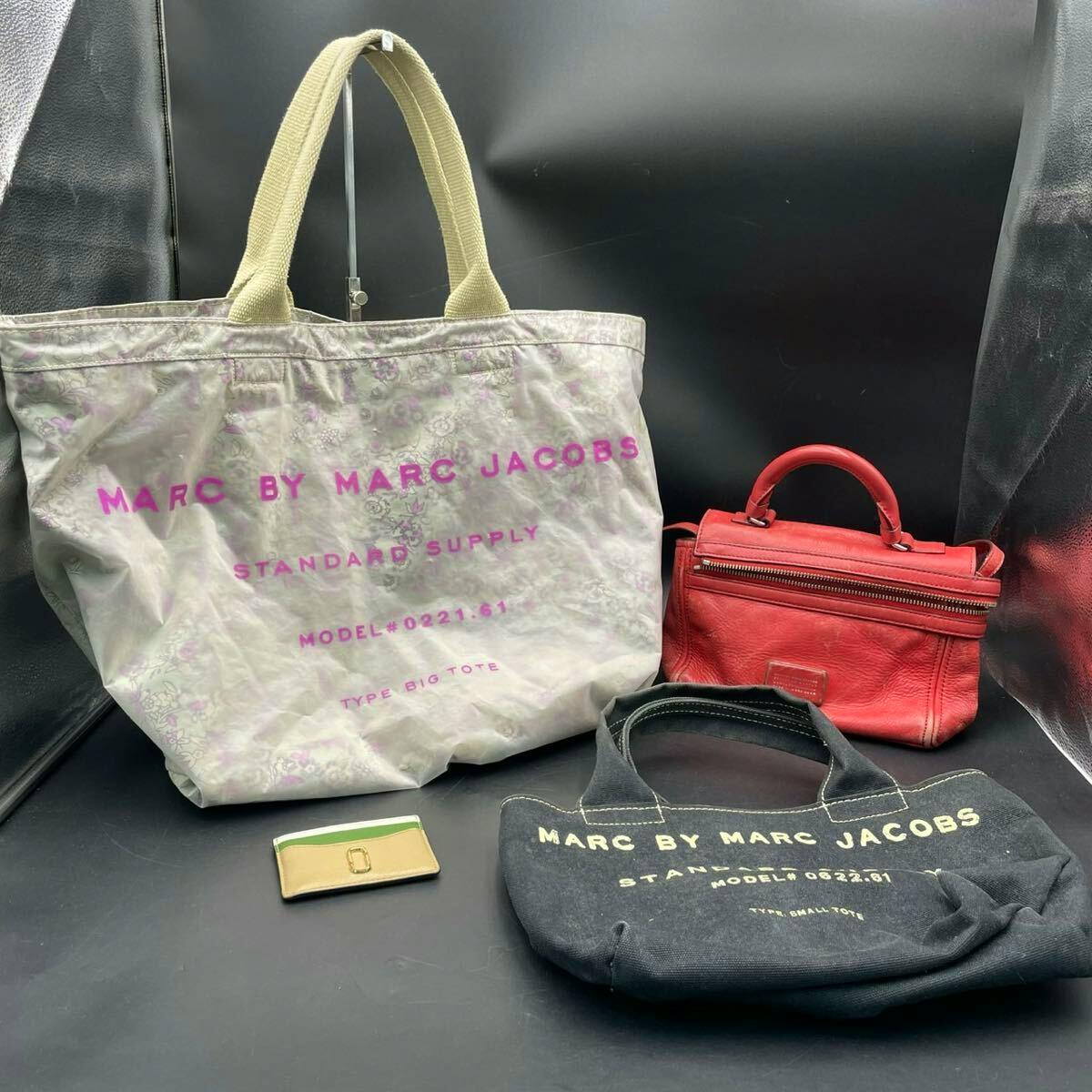 Four Marc by Marc Jacobs items: a large printed tote bag, a small black tote, a red handbag, and a card case arranged on a black background.