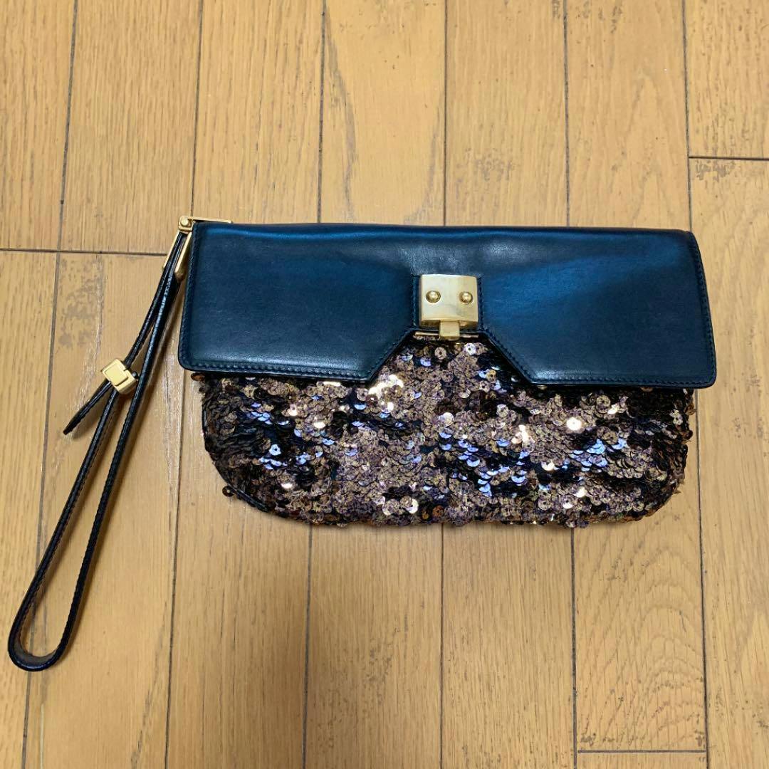 A MARC JACOBS clutch bag with a black leather flap, gold clasp, sequin body, and attached wrist strap on a wooden floor.