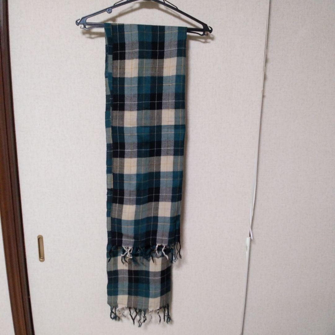 Checked wool muffler with teal, black, and cream plaid pattern hanging from a hanger against a light wall.