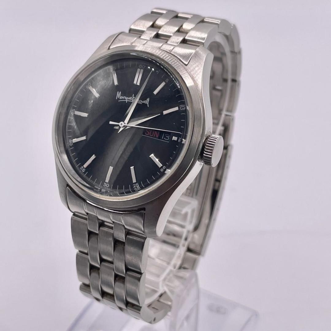 MargaretHowell men's automatic day-date wristwatch with a black dial and metal bracelet shown on a display stand.