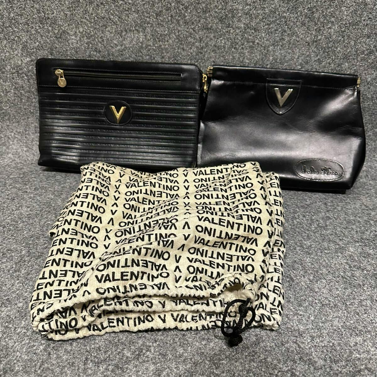 Two black Valentino leather clutch bags placed behind a folded Valentino dust bag on a gray surface.