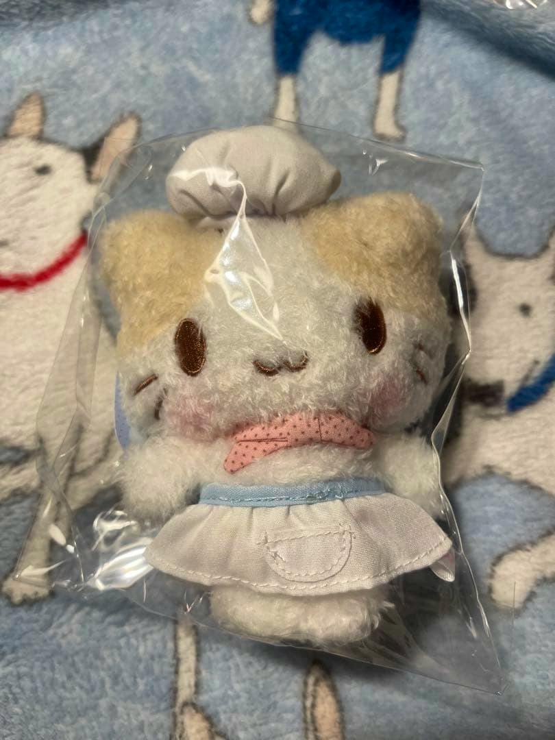 Front view of a marshmallow-like fluffy cat plush mascot wearing a chef outfit, shown inside clear plastic packaging.