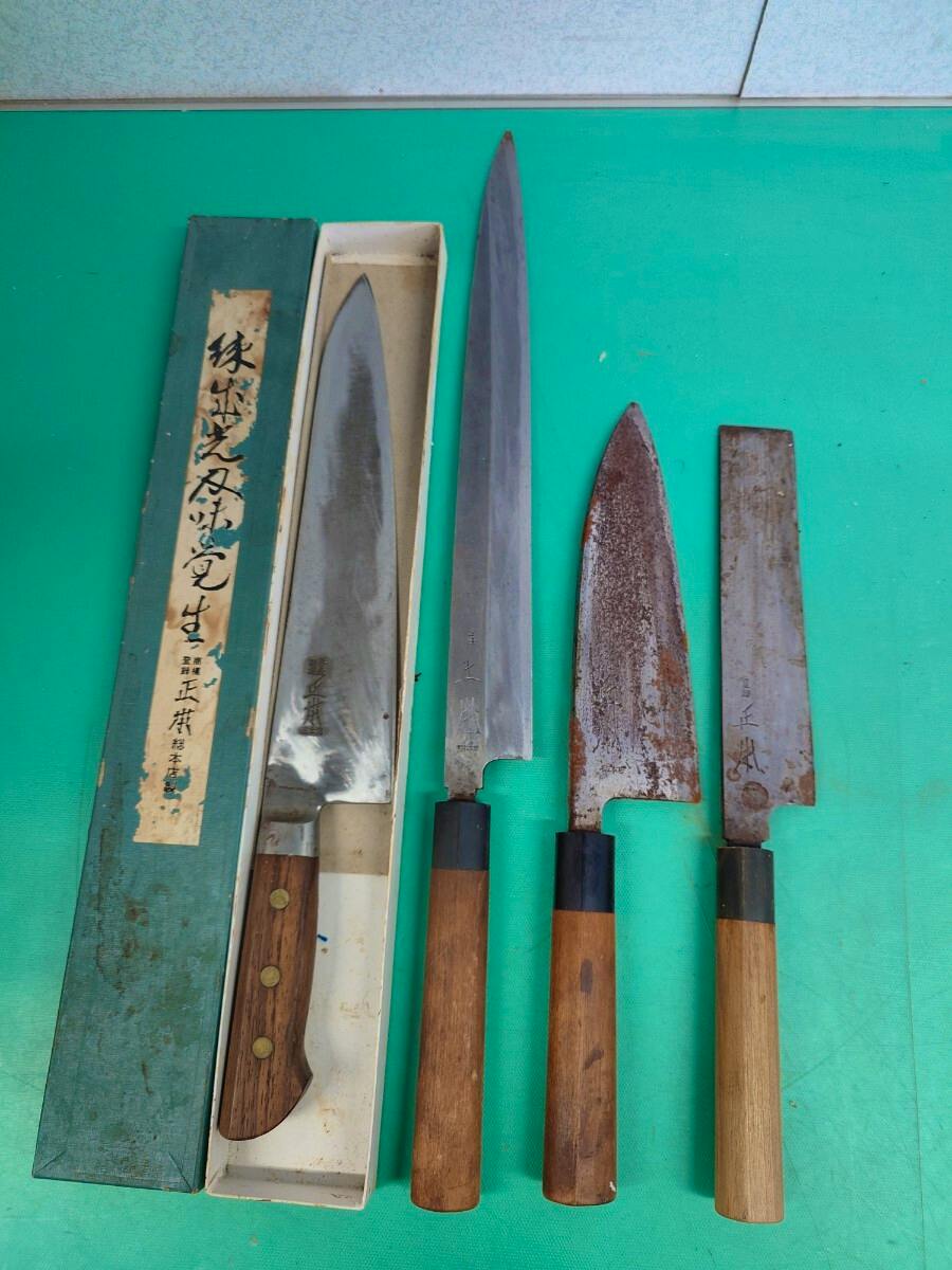 Four used chef knives with wooden handles laid out on a green mat next to a green box, showing blades and handles.