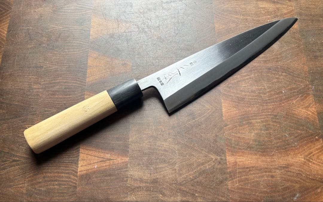 Full Japanese deba-style chef knife with a light wooden handle and engraved steel blade lying on a wooden cutting board.