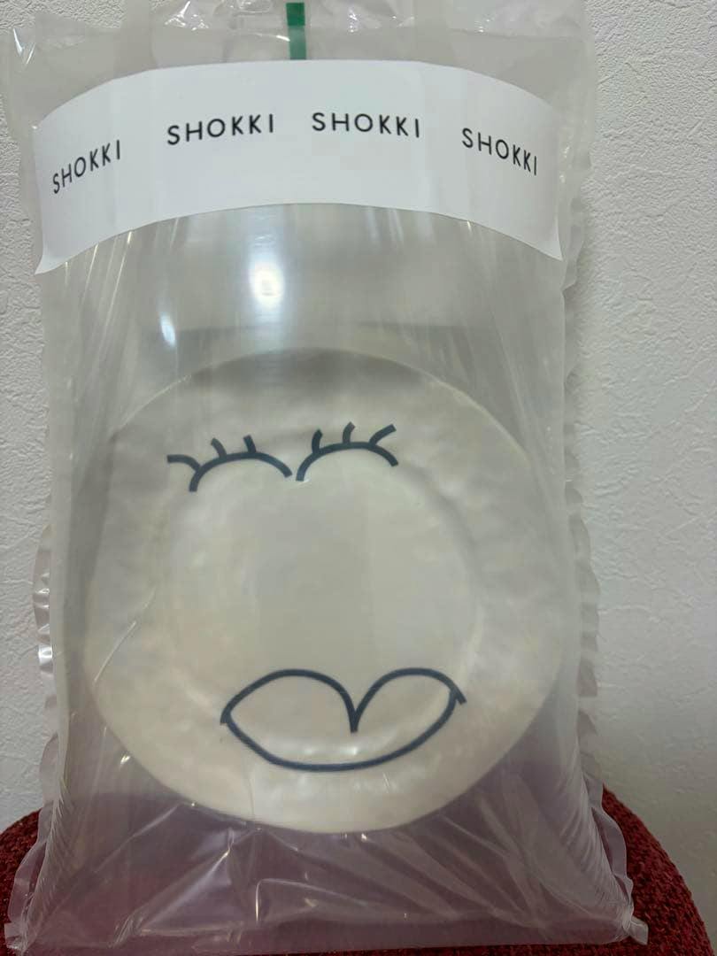 Front of a SHOKKI ceramic plate with a simple face motif visible through clear plastic packaging.