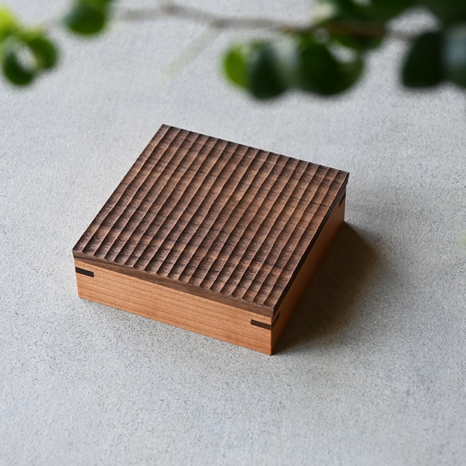 A closed square wooden box with a carved textured lid resting on a light surface.