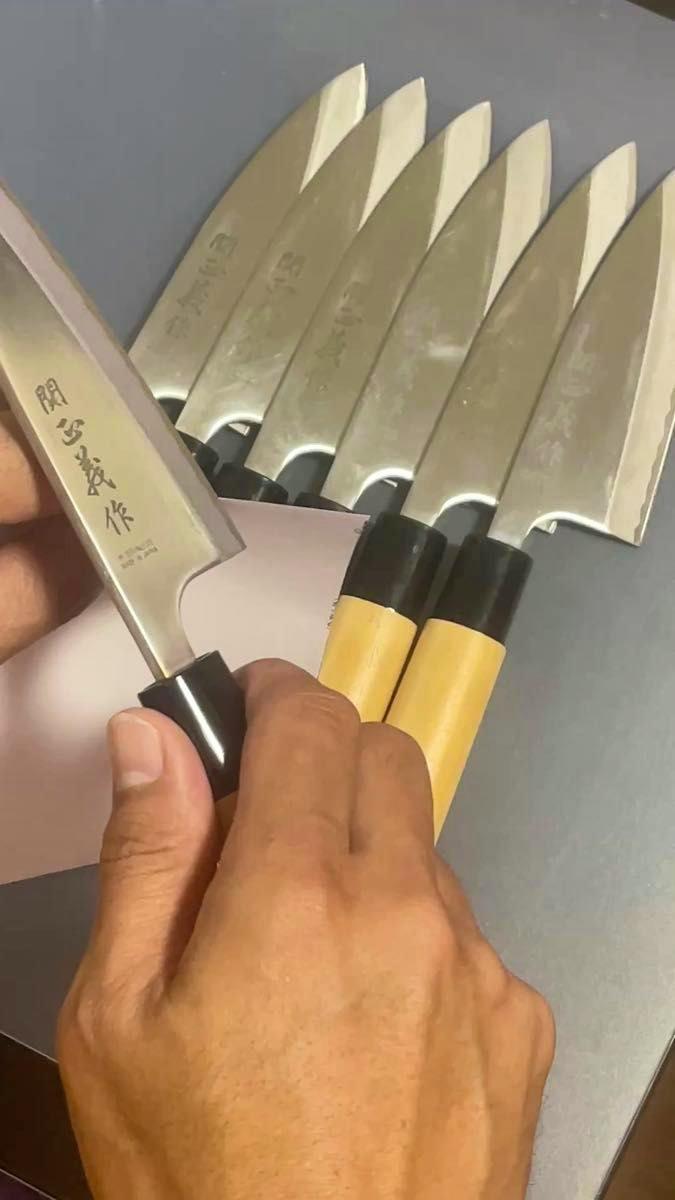 A hand holding a 関正義作 出刃包丁 (deba knife) with several similar knives lined up showing blades and wooden handles.