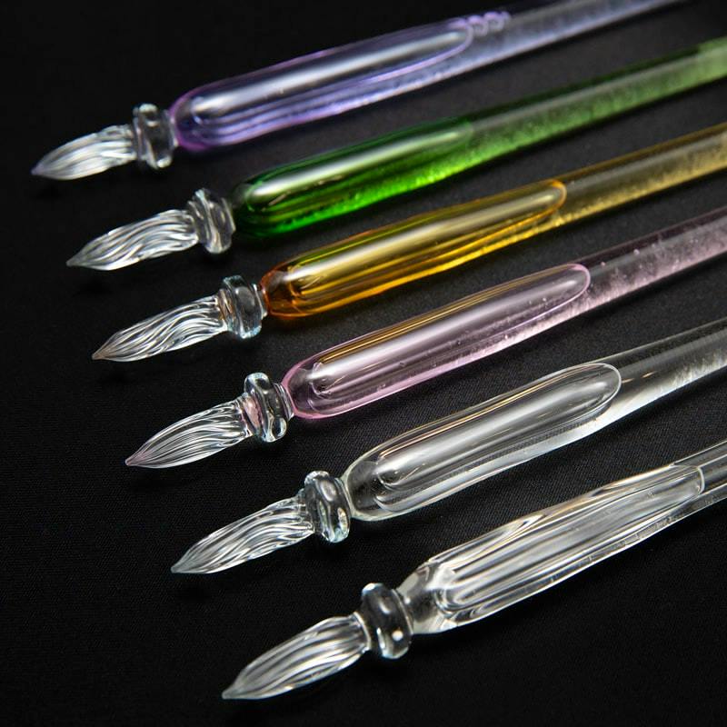Six glass fountain (dip) pens in various colors arranged on a black surface.