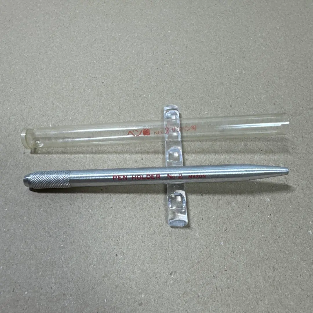 Silver metal pen holder (pen shaft) with clear plastic case on a plain cardboard background.