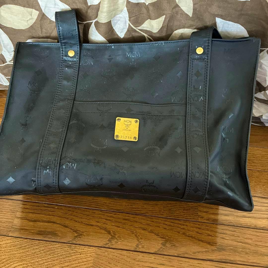 Black MCM canvas tote bag with shoulder straps and a gold logo plate on the front, shown lying on a wooden floor.