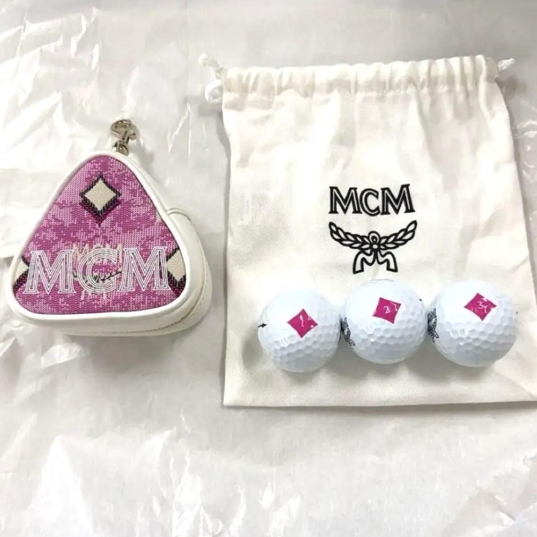 A triangular pink MCM pouch, three white golf balls with pink square logos, and a white MCM drawstring bag on a white surface.