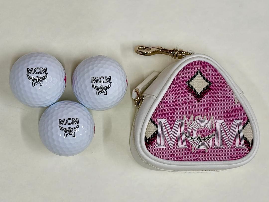 Three white MCM golf balls and a pink triangular MCM golf ball pouch with white trim and a gold clasp on a light background.