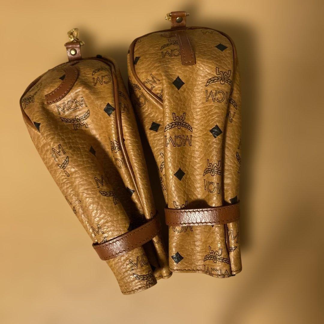 Two brown MCM-pattern golf club head covers placed on a tan background.