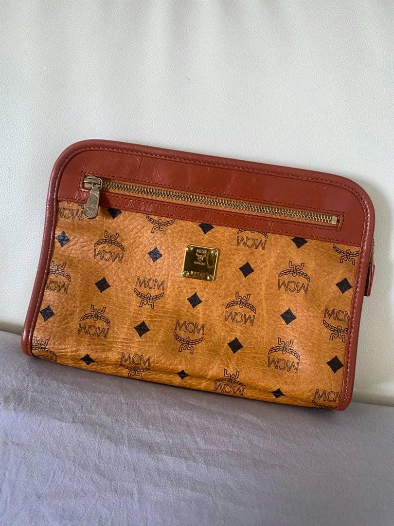 Brown MCM logo-print leather clutch bag showing a front zip pocket and a gold logo plate, resting against a white background.