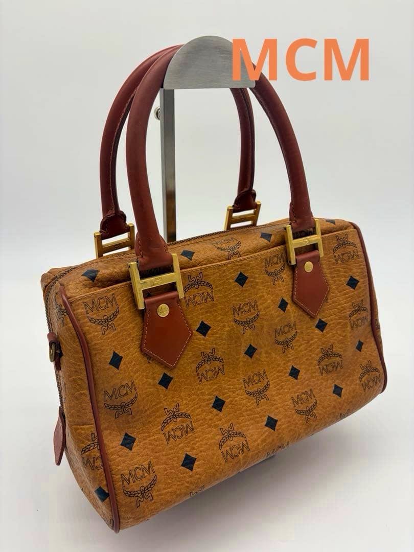 Brown MCM mini Boston handbag with monogram pattern, dual top handles, and gold hardware displayed on a stand.