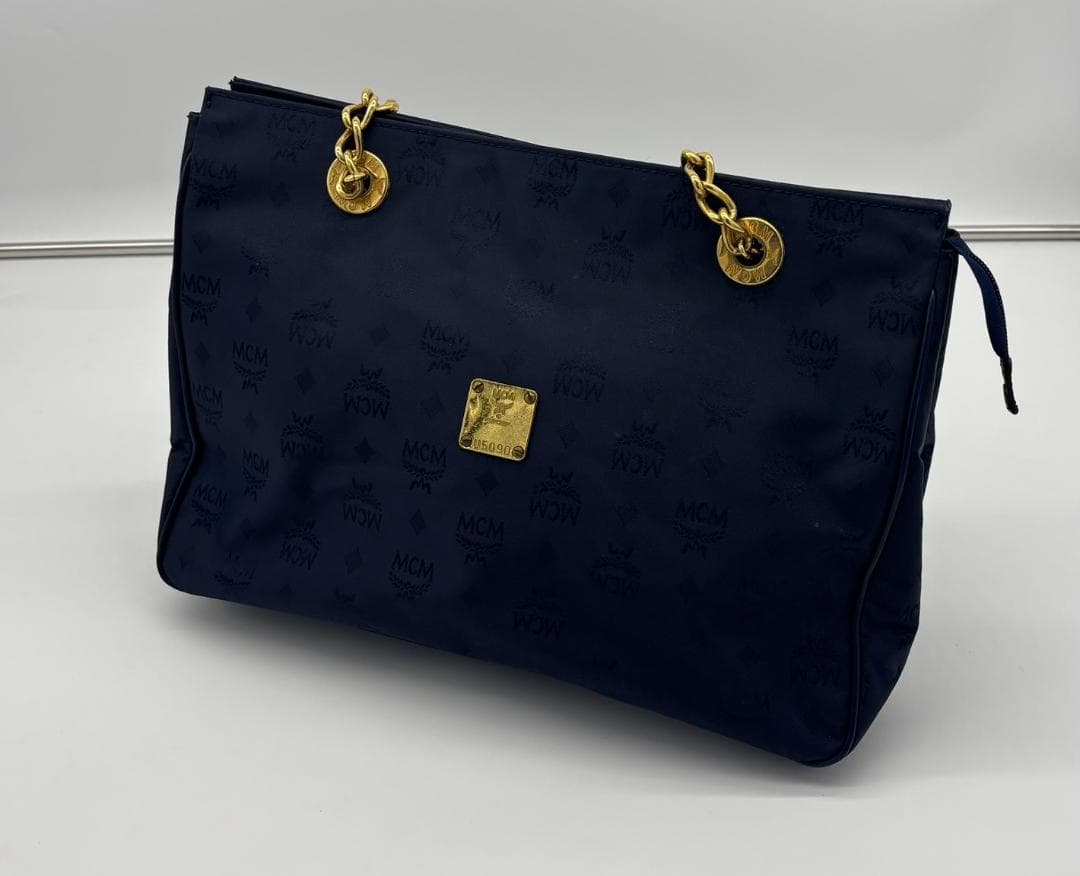 Dark blue MCM monogram Visetos-pattern chain-shoulder tote bag with gold-tone hardware and front logo plaque on a white background.