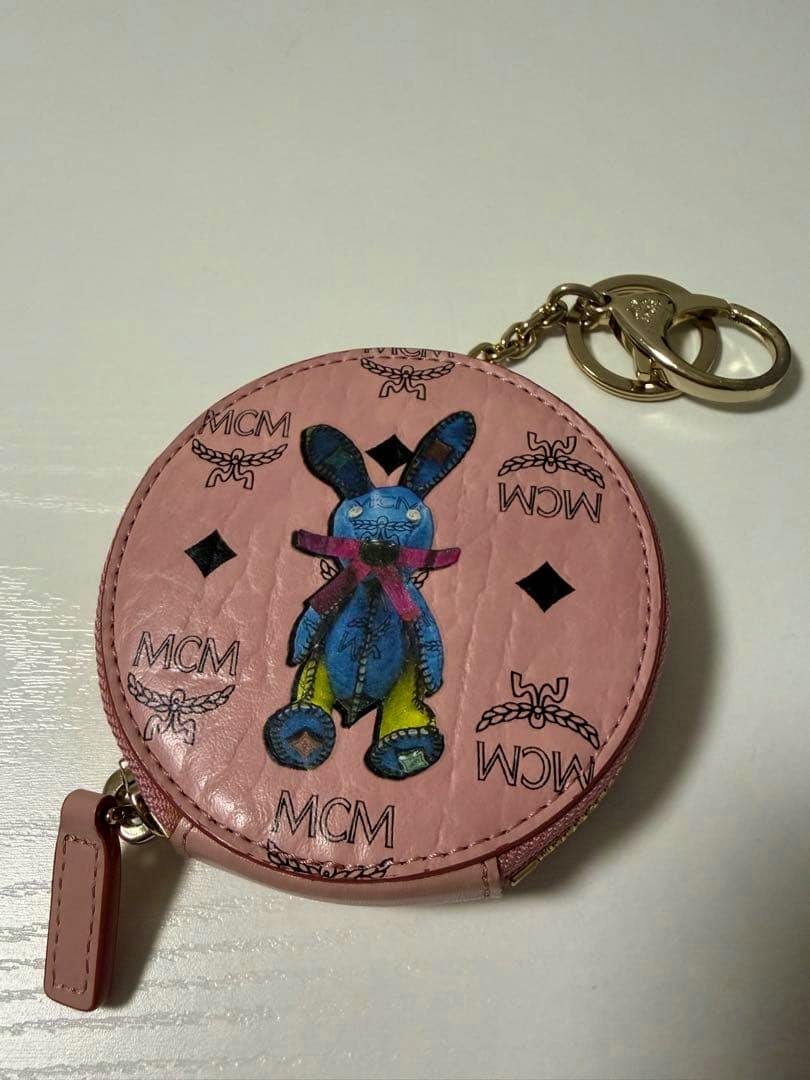 Pink round MCM coin case key holder in cow leather with MCM monogram and a blue rabbit illustration, shown with a gold keyring.