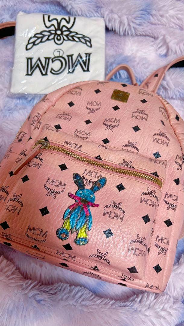 Pink MCM backpack with repeating MCM monogram and a colorful rabbit illustration on the front pocket, shown laying on a plush surface with a folded MCM dust bag visible.