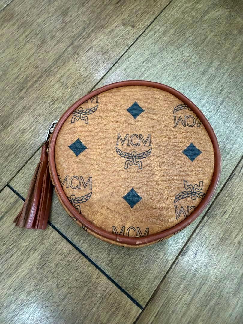 Round brown MCM leather coin case with printed logo pattern and a tassel, photographed on a wooden floor.