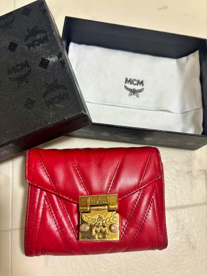 Red MCM tri-fold wallet with a gold clasp displayed in front of its black MCM box and white dust bag.