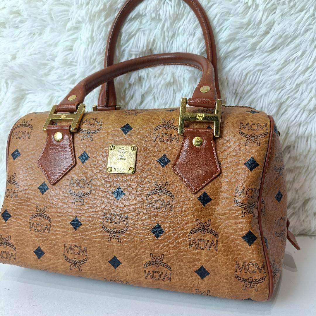 Tan MCM Visetos PVC vintage Boston handbag with brown handles, gold hardware, and logo monogram on a white background.