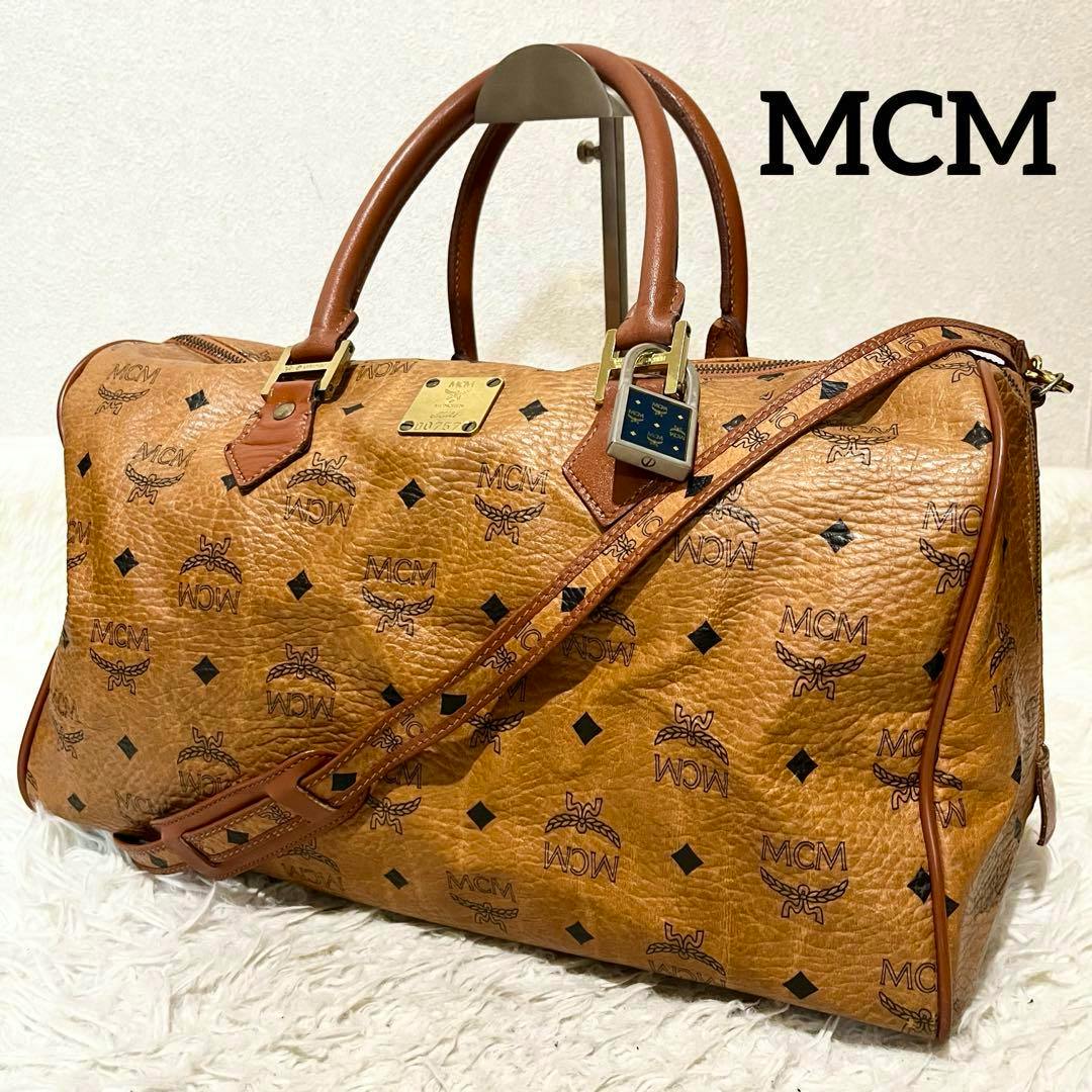 Brown MCM canvas Boston bag with all-over logo print, top handles, a shoulder strap, and a padlock on the zipper.