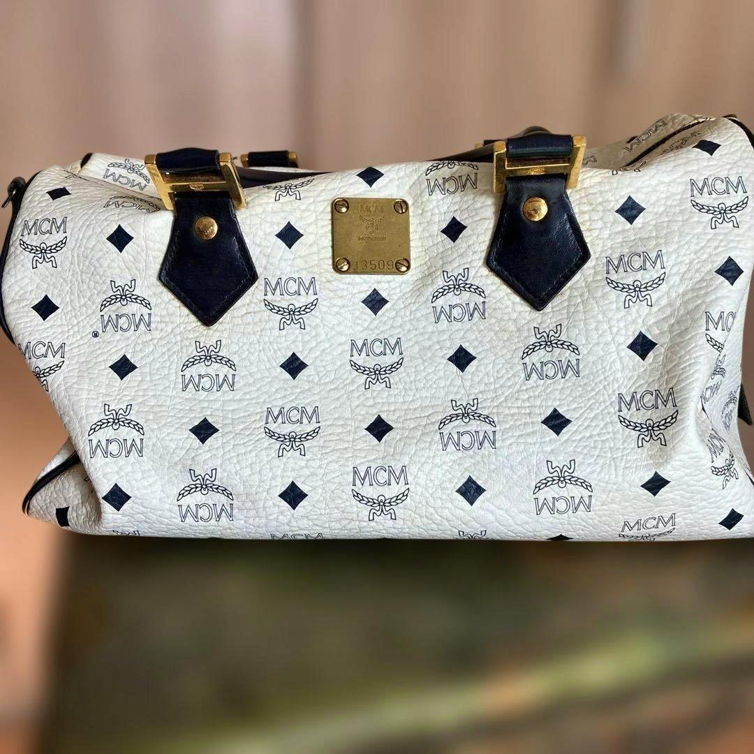 White MCM Visetos Boston bag showing the black repeated MCM logo pattern, black leather handle attachments, and a central gold logo plate.