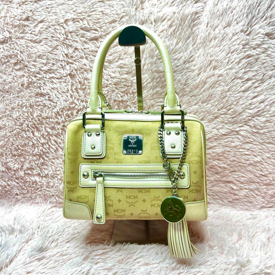 Beige MCM Visetos mini handbag with front zipper pocket, tassel charm, and double handles on a pink fuzzy background.