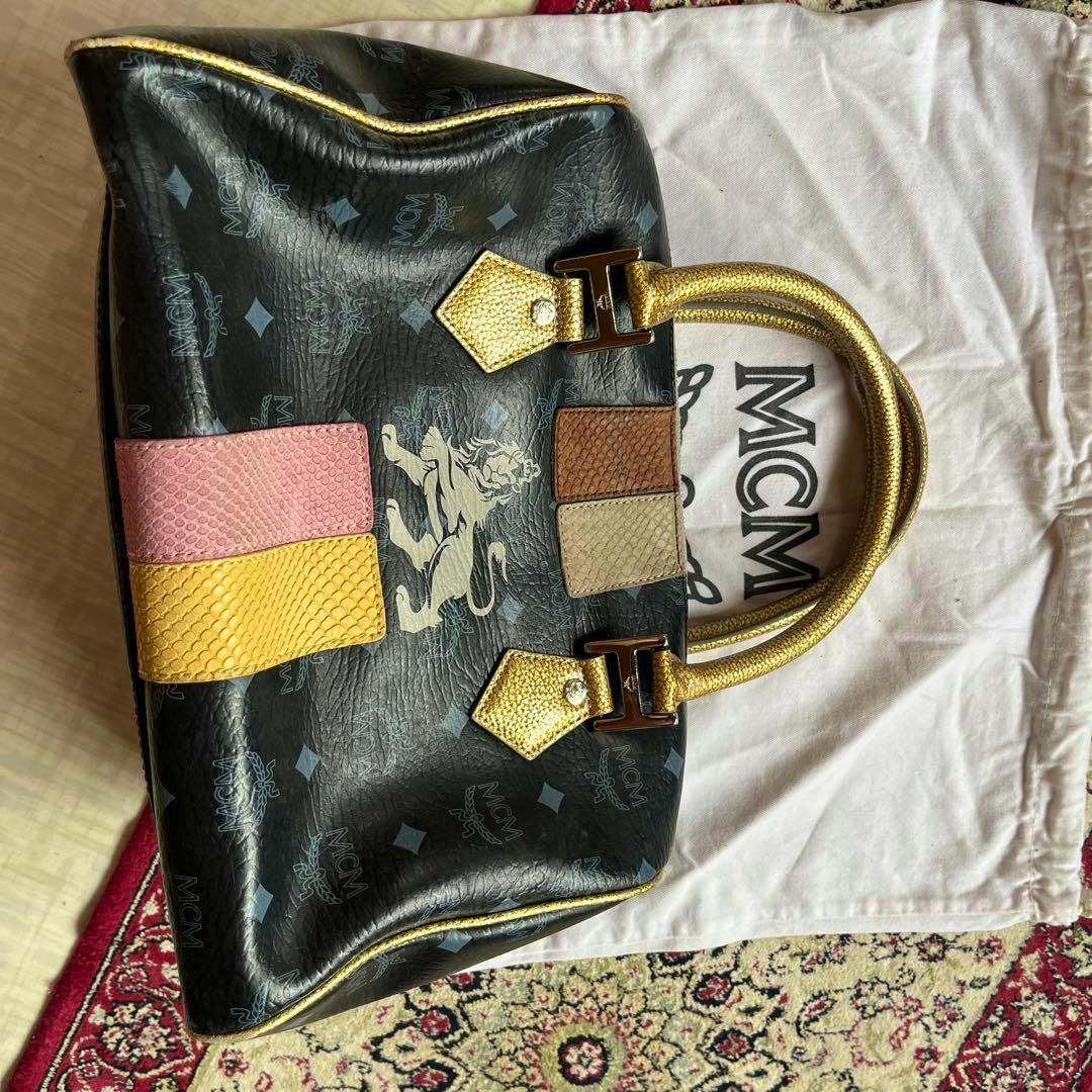 Black MCM Visetos-pattern canvas handbag with gold handles and colored patch accents placed on a white dust bag.
