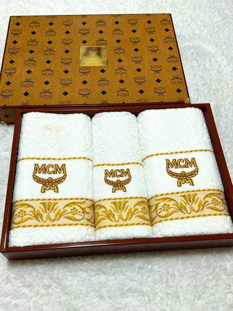 Three white MCM-branded towels (one wash towel and two face towels) arranged in a presentation box showing gold embroidery and patterned border.