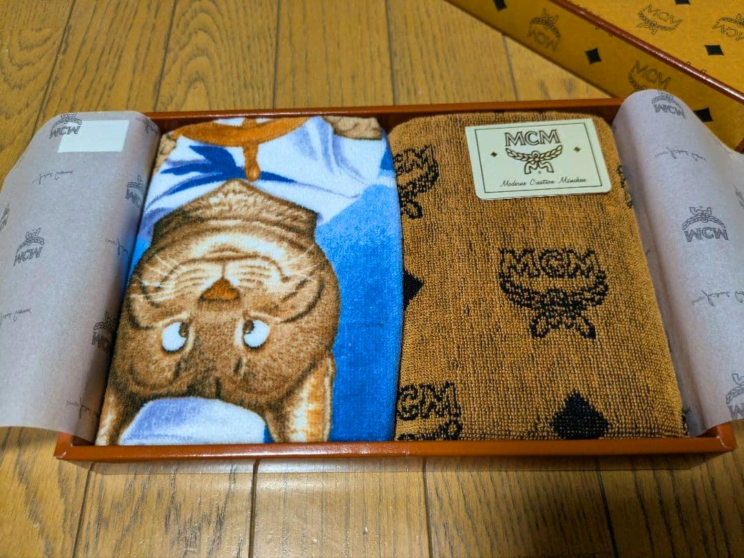 Two MCM wash towels in an open branded box, one brown MCM logo towel and one blue towel with a cat face.
