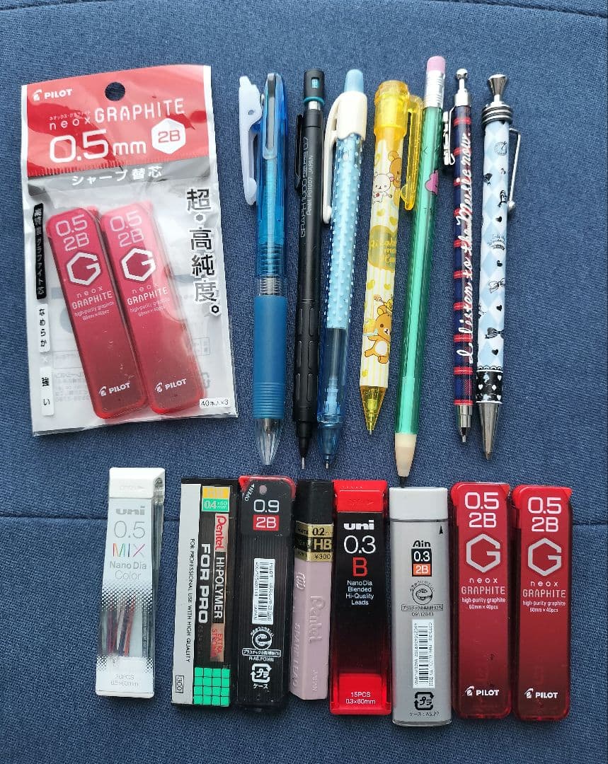 Photo showing multiple mechanical pencils and several tubes of mechanical pencil lead refills arranged on blue fabric.