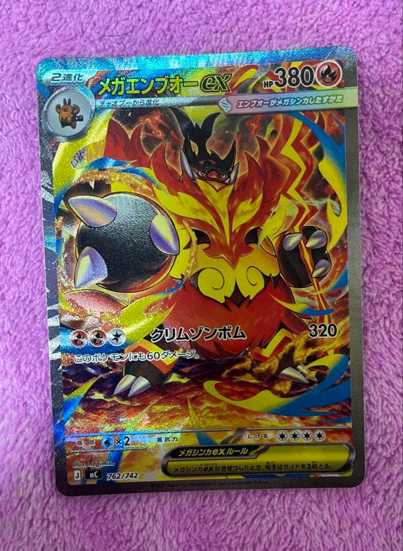 Photograph of the trading card メガエンブオーex showing the full holographic card on a pink towel background.