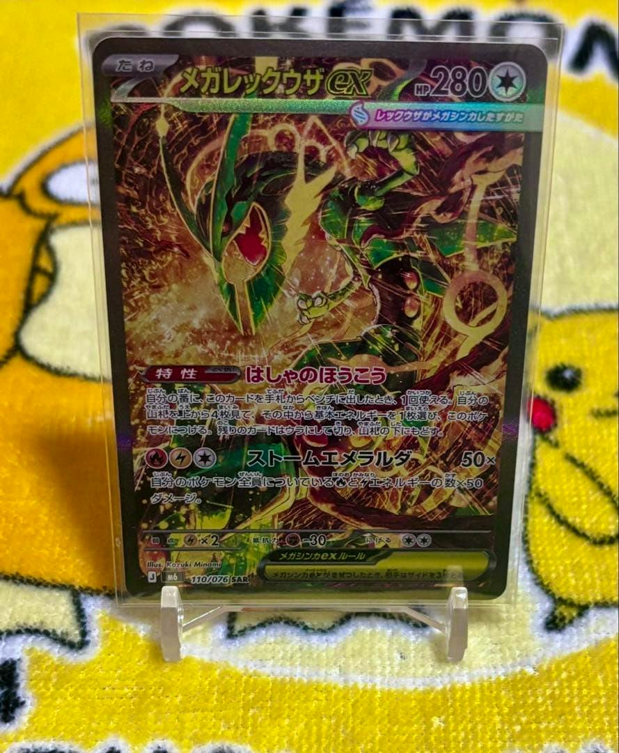 Photograph of the メガレックウザex trading card in a clear protective sleeve standing on a yellow Pikachu towel.