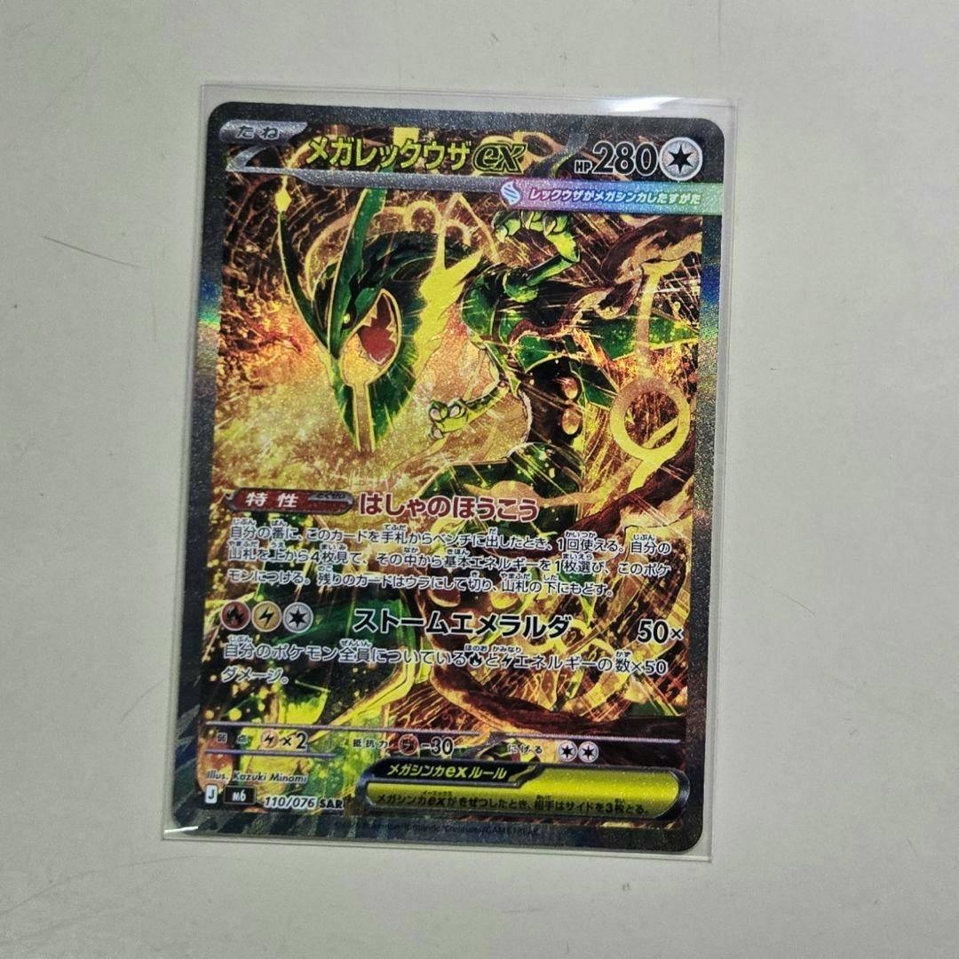 Photograph of the メガレックウザex SAR trading card in a clear protective sleeve on a light surface.
