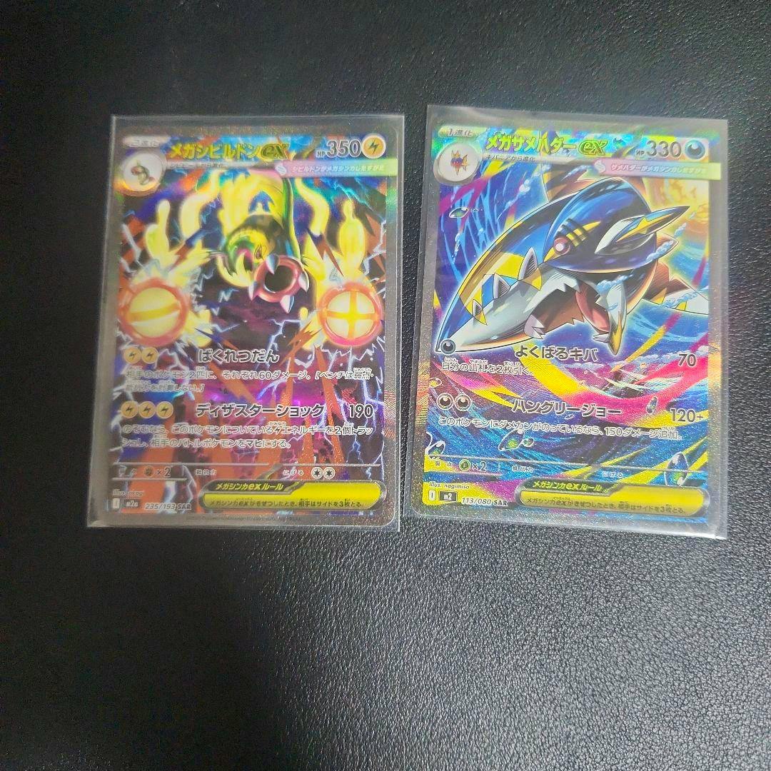 Two holographic trading cards from the メガサメハダーex / メガシビルドンex SAR two-card set in protective sleeves placed side by side on a black surface.