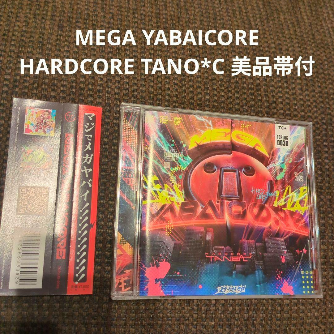 Closed CD jewel case of MEGA YABAICORE HARDCORE TANO*C showing the album front cover and the separate red obi strip on a brown carpeted background, with white promotional text at the top.