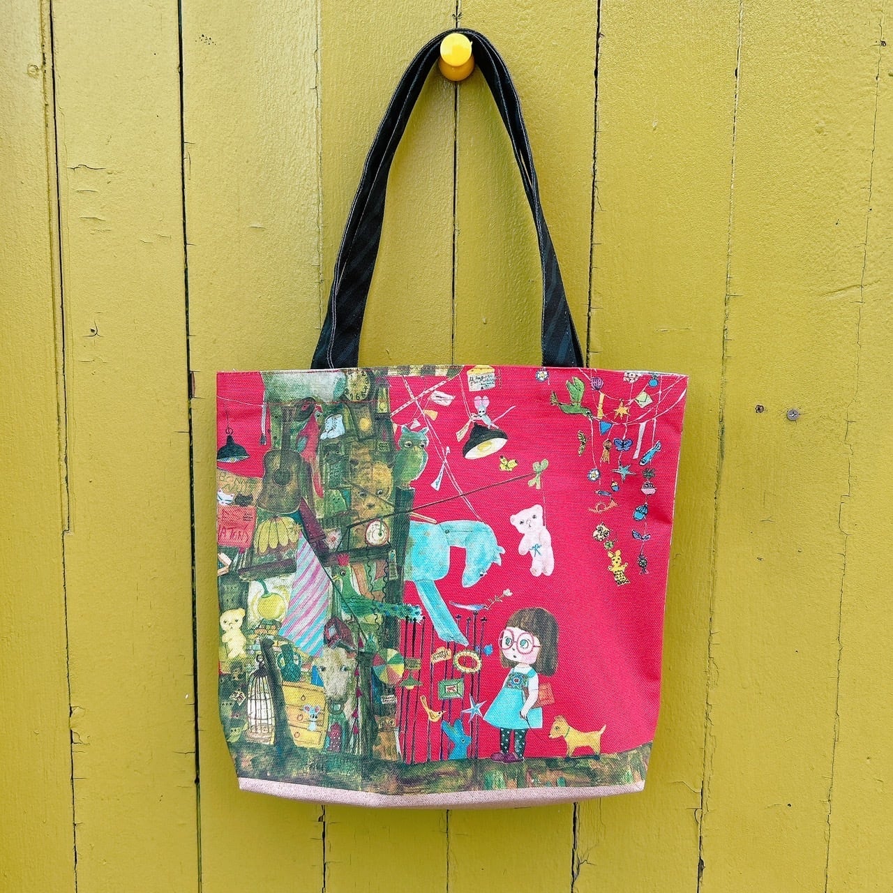 Reusable tote bag with a colorful illustration of a girl wearing glasses and a horse on a red background, hanging by black handles on a yellow wall.