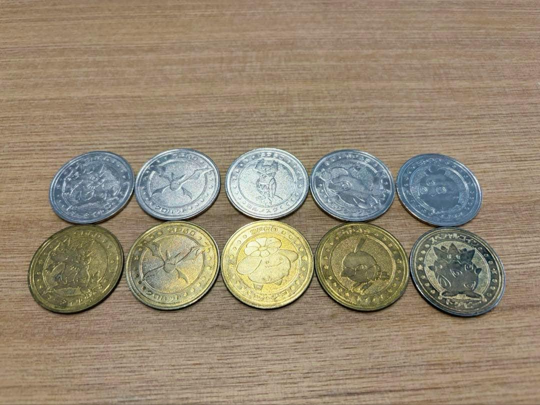 Ten metal Pokémon battle coins arranged in two rows on a wooden surface showing character designs on the coin faces.